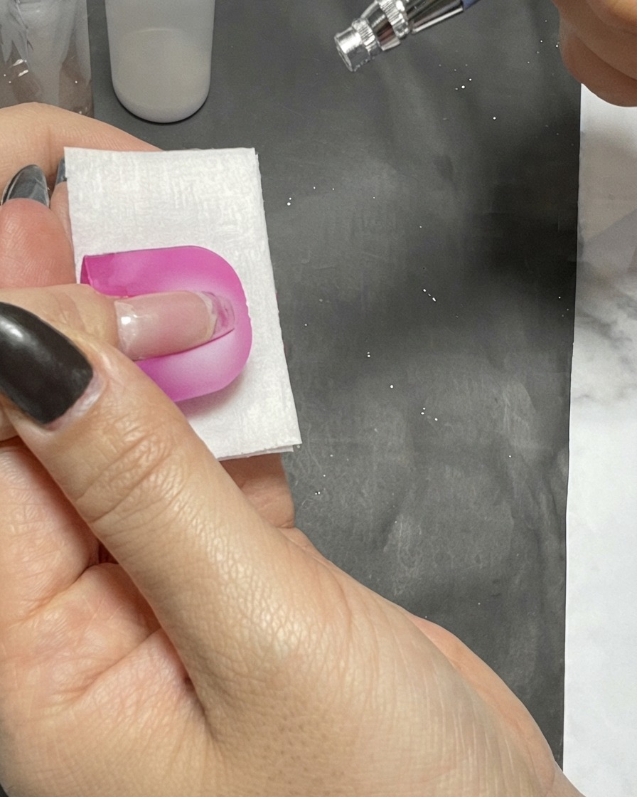 Un stylo aérographe vaporisant une fine brume sur un ongle protégé par une barrière rose peel-off.