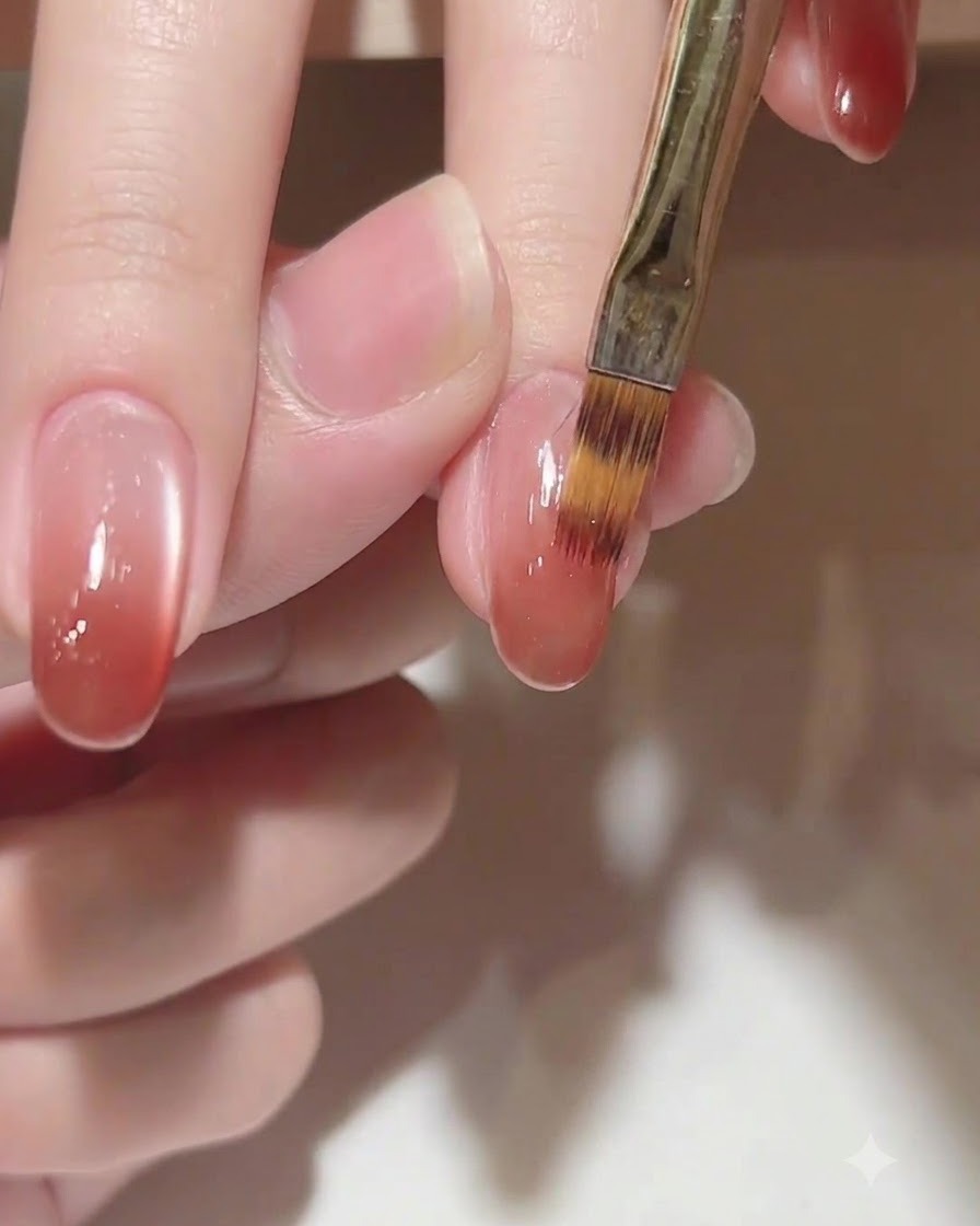 Gros plan sur un pinceau estompant délicatement un dégradé de couleur gel brun-rosé chaleureux vers les cuticules sur un ongle oval.