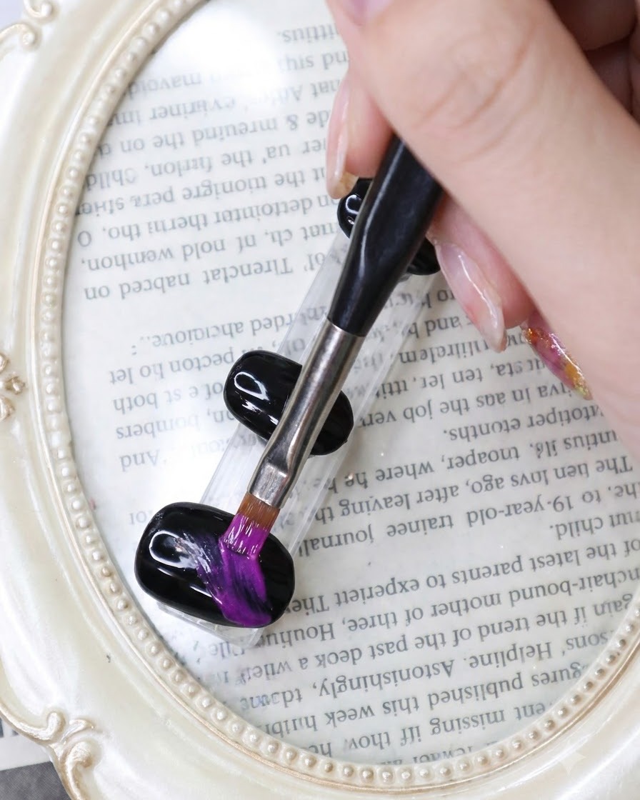 Un pinceau fin traçant des traits vibrants en violet néon à travers un ongle en gel noir sur un fond sur le thème des livres.