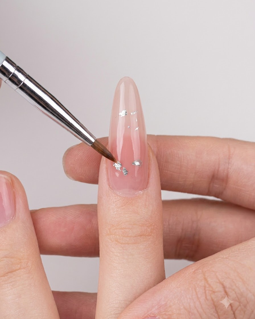Un pinceau de nail art fin déposant de petits flocons iridescents argentés sur un ongle nude brillant.