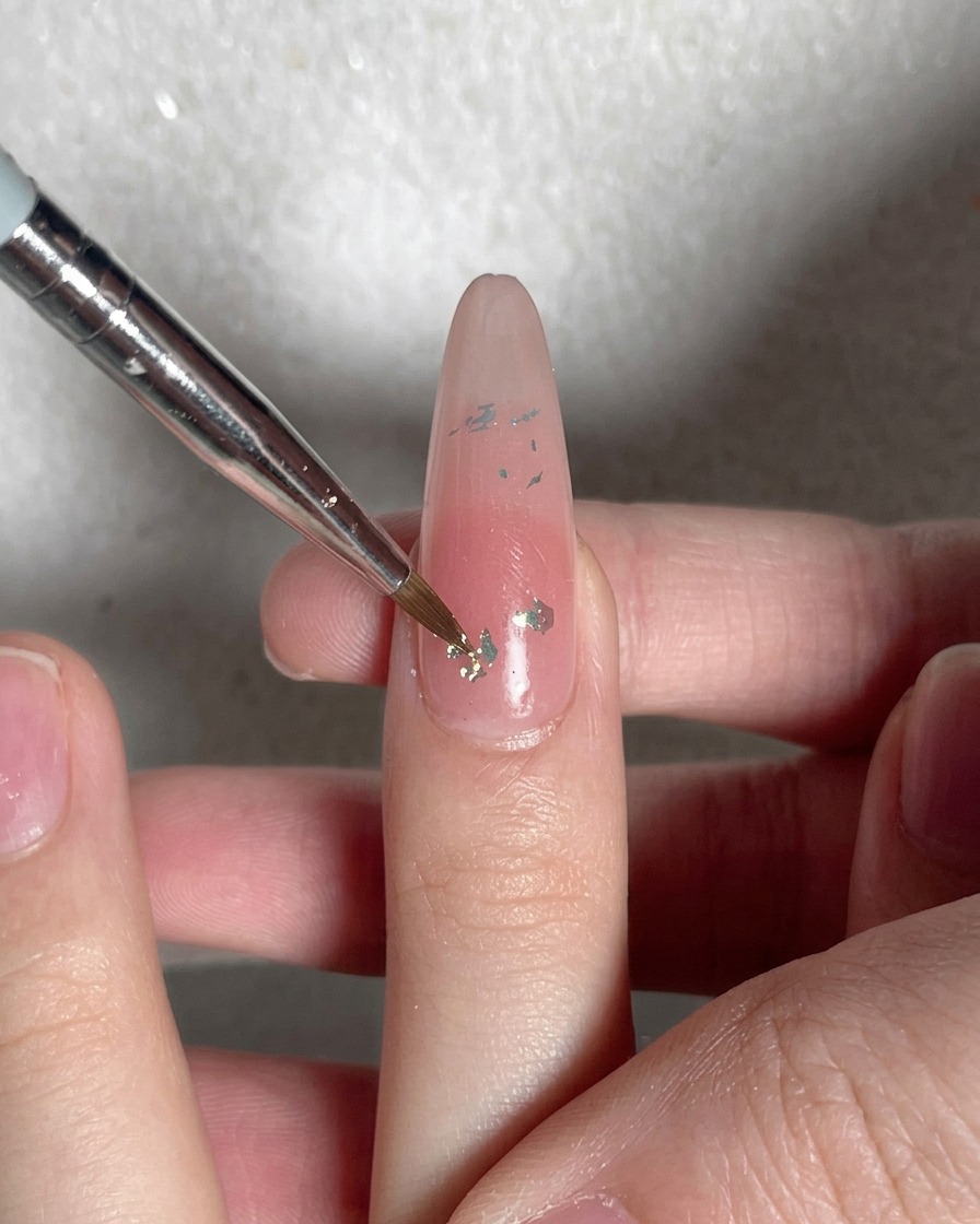 Un gros plan sur la pointe dun pinceau pressant à plat des flocons iridescents sur un ongle nude.