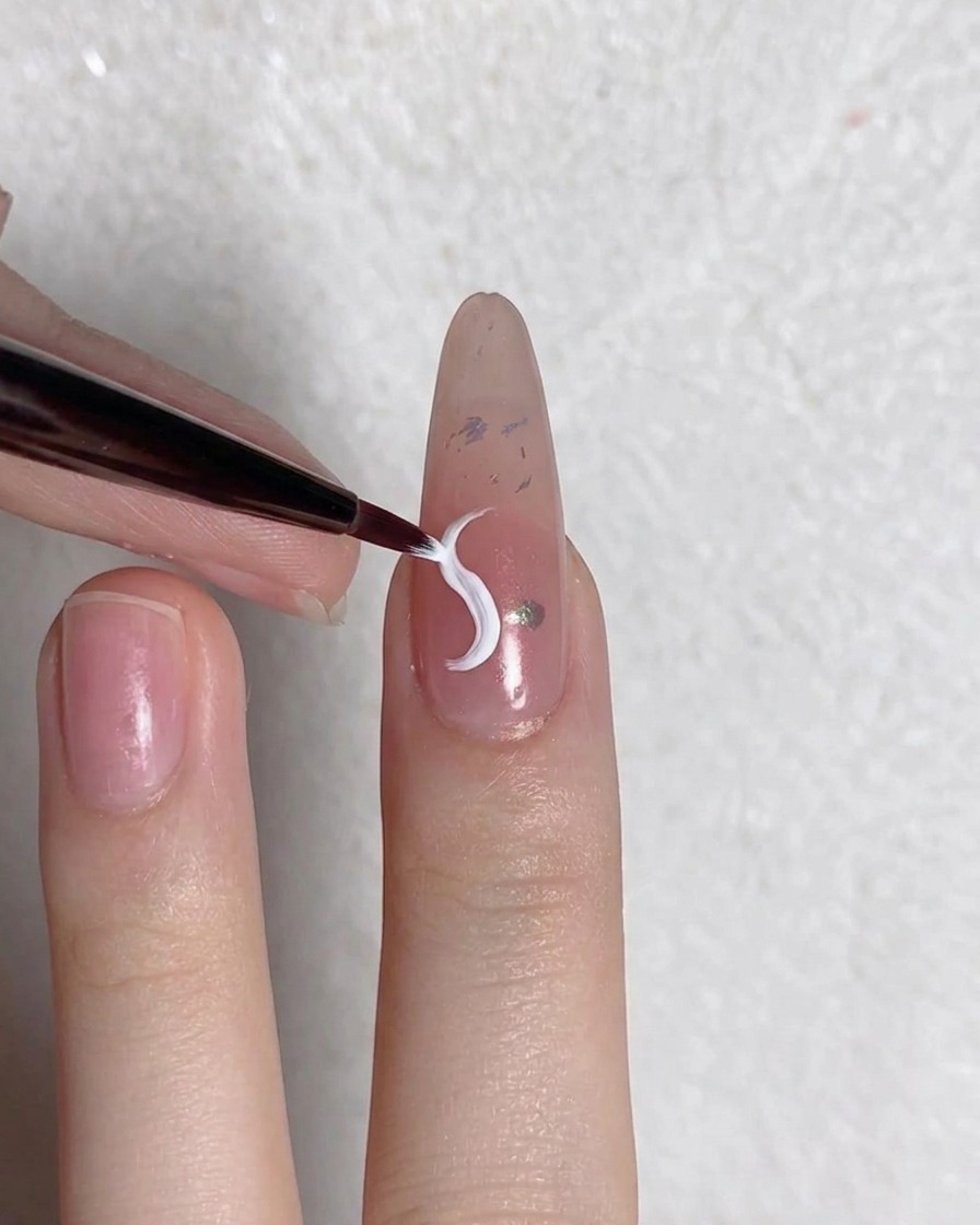 Un pinceau fin trace une ligne de pétale blanche incurvée sur un ongle préparé.