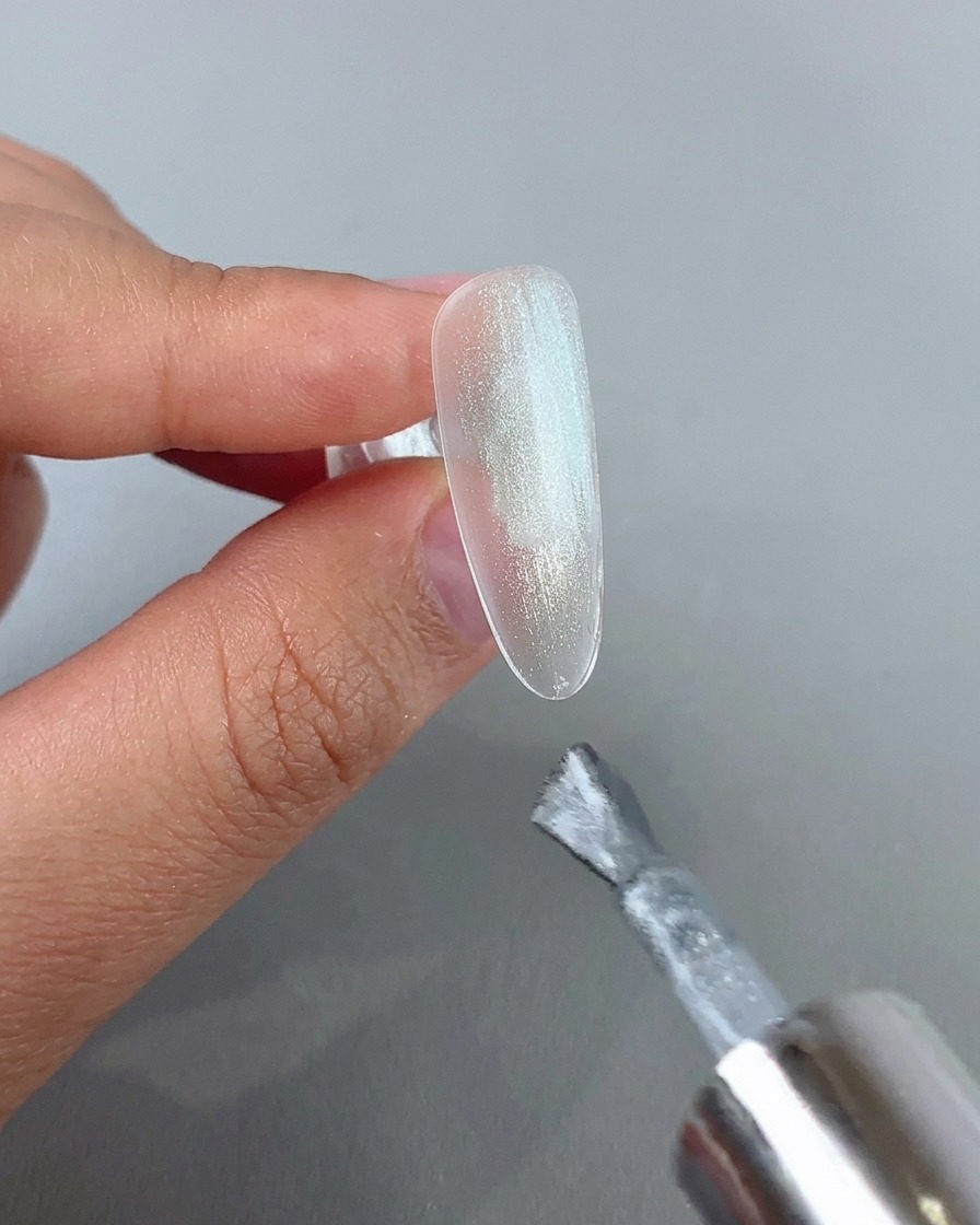 Un pinceau à ongles appliquant une couche de gel cat-eye argenté scintillant sur une capsule dongle transparente.