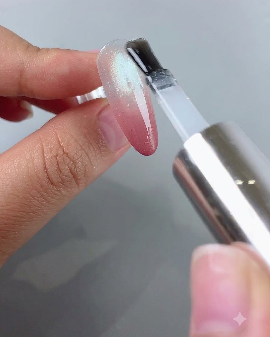 Un pinceau appliquant un top coat transparent sur un ongle doté dun blush dégradé rose et dun effet cat-eye argenté.