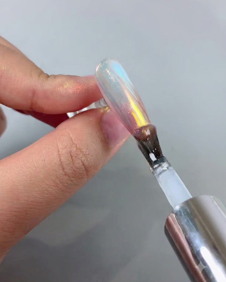 Un pinceau à ongles appliquant un top coat transparent sur un ongle doté dune finition chrome aurora du rose au bleu.