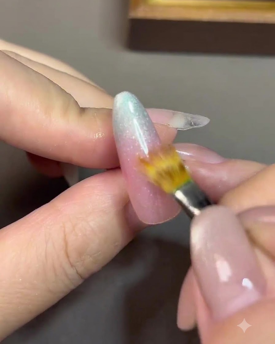 Un pinceau de détail fin estompant la ligne de transition entre un gel rose nude et un gel bleu sur un ongle.