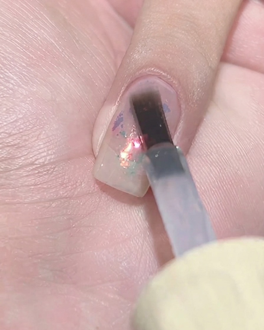 Un pinceau plat pour gel tapotant une couche de vernis violet transparent sur une grappe de paillettes texturée sur un onglet.
