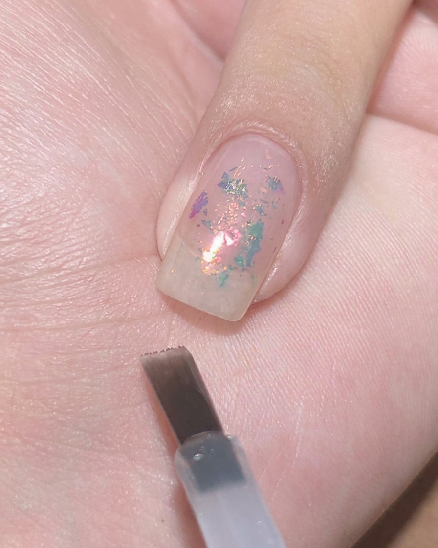 Un pinceau gel plat surplombant un ongle transparent avec des paillettes incrustées, prêt à tapoter du vernis violet diaphane.