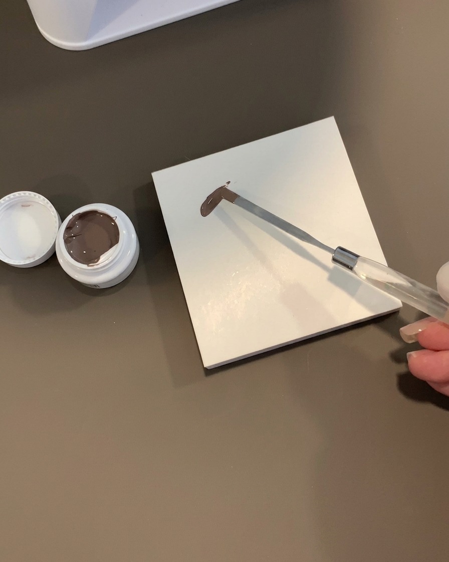 Une spatule métallique tenant une noisette de gel gris-marron sur une palette en carreau blanc carré à côté dun pot ouvert.