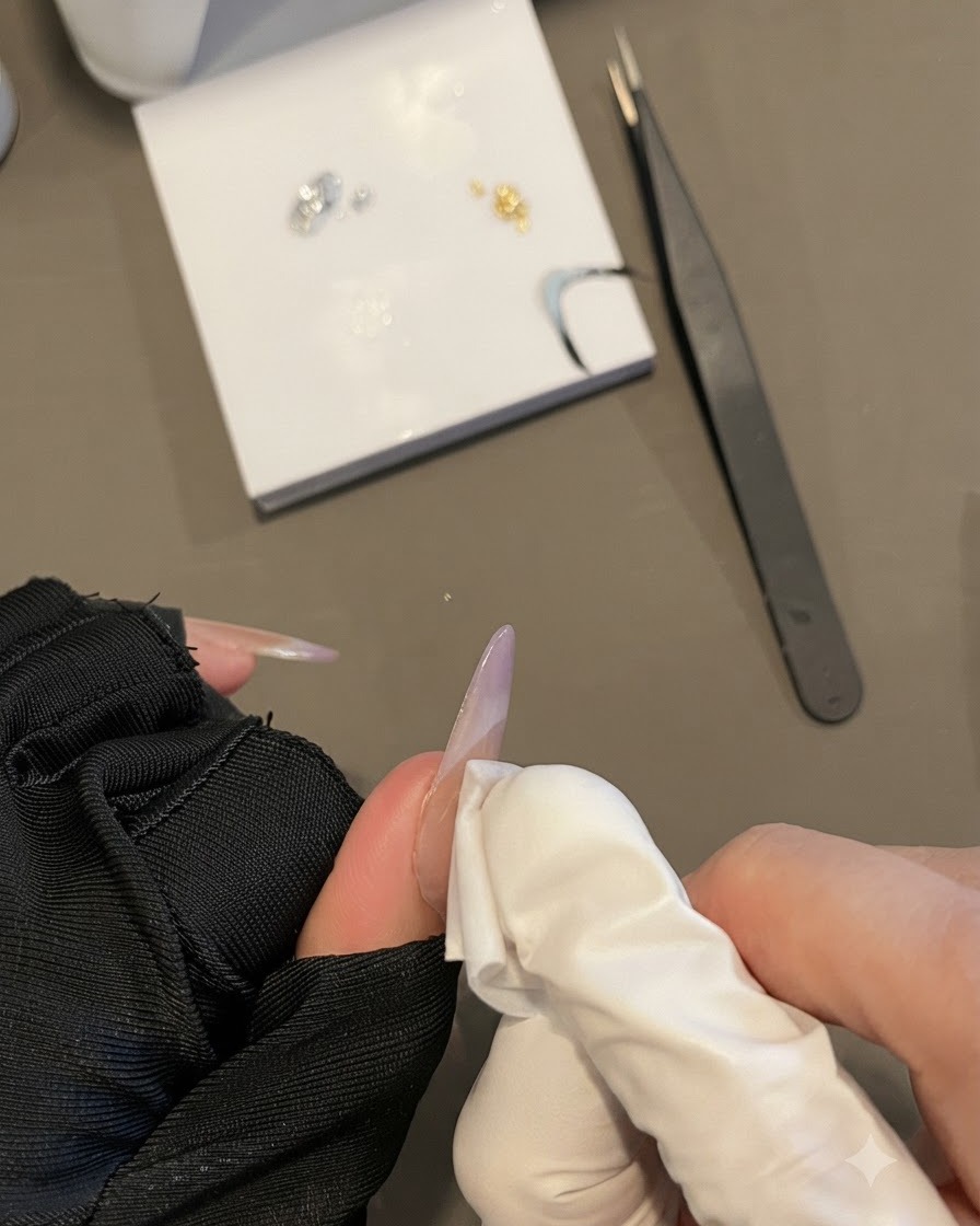 Une main gantée essuyant la surface dun ongle avec une lingette non pelucheuse tout en tenant un long ongle ovale.