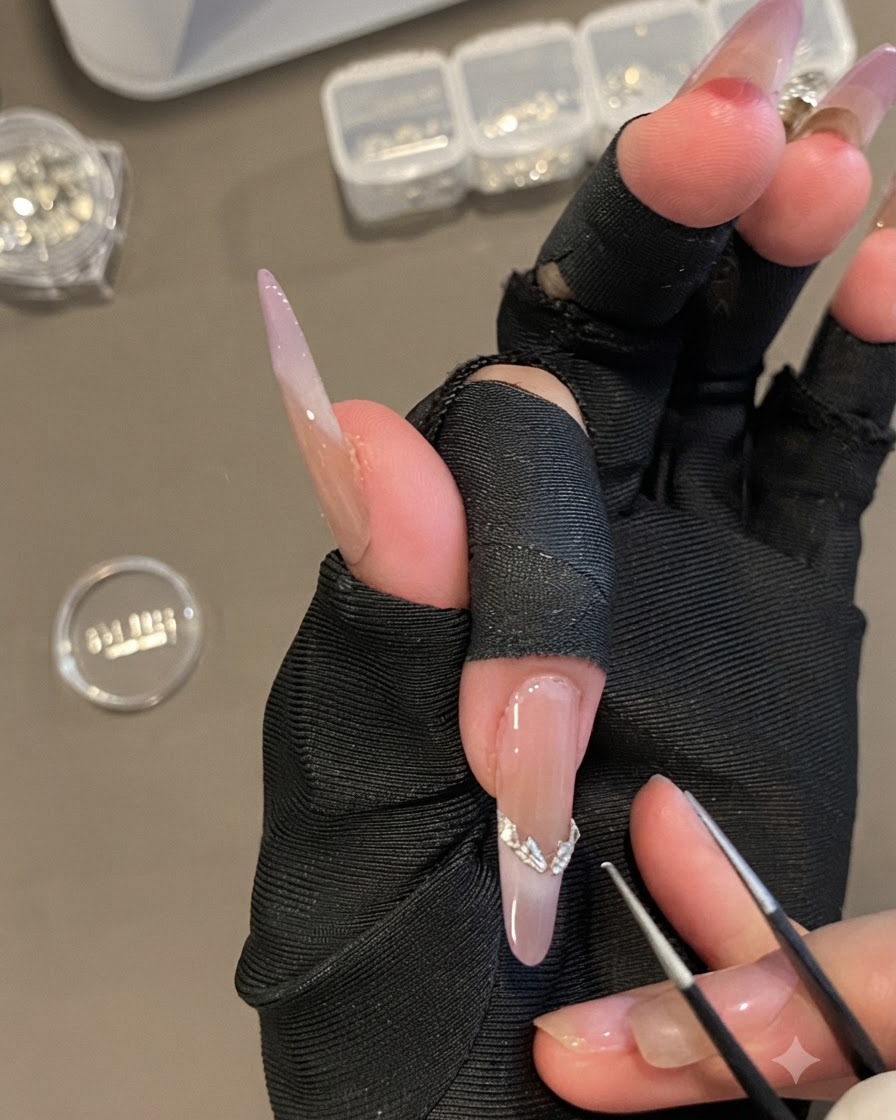 Une main tenant une pince fine pour placer un autocollant courbé argenté de French tip sur un long ongle ovale.