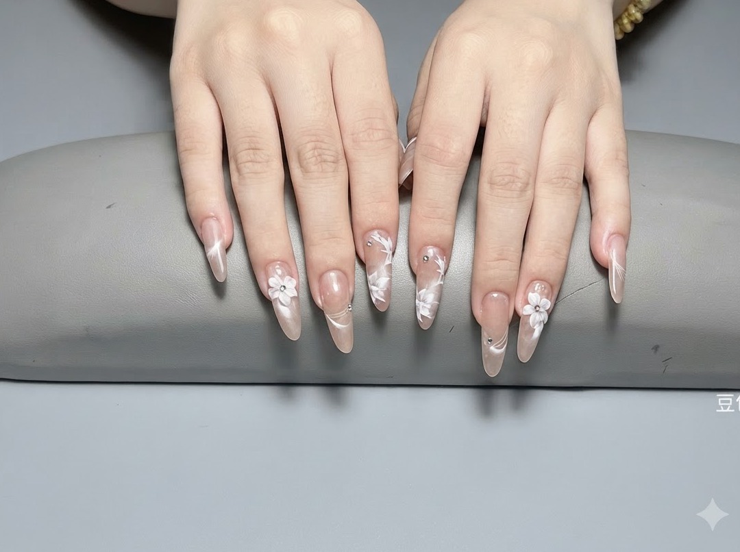 Gros plan sur des ongles longs en amande ornés de fleurs blanches 3D sculptées et de minuscules cristaux argentés sur une base nude brillante.