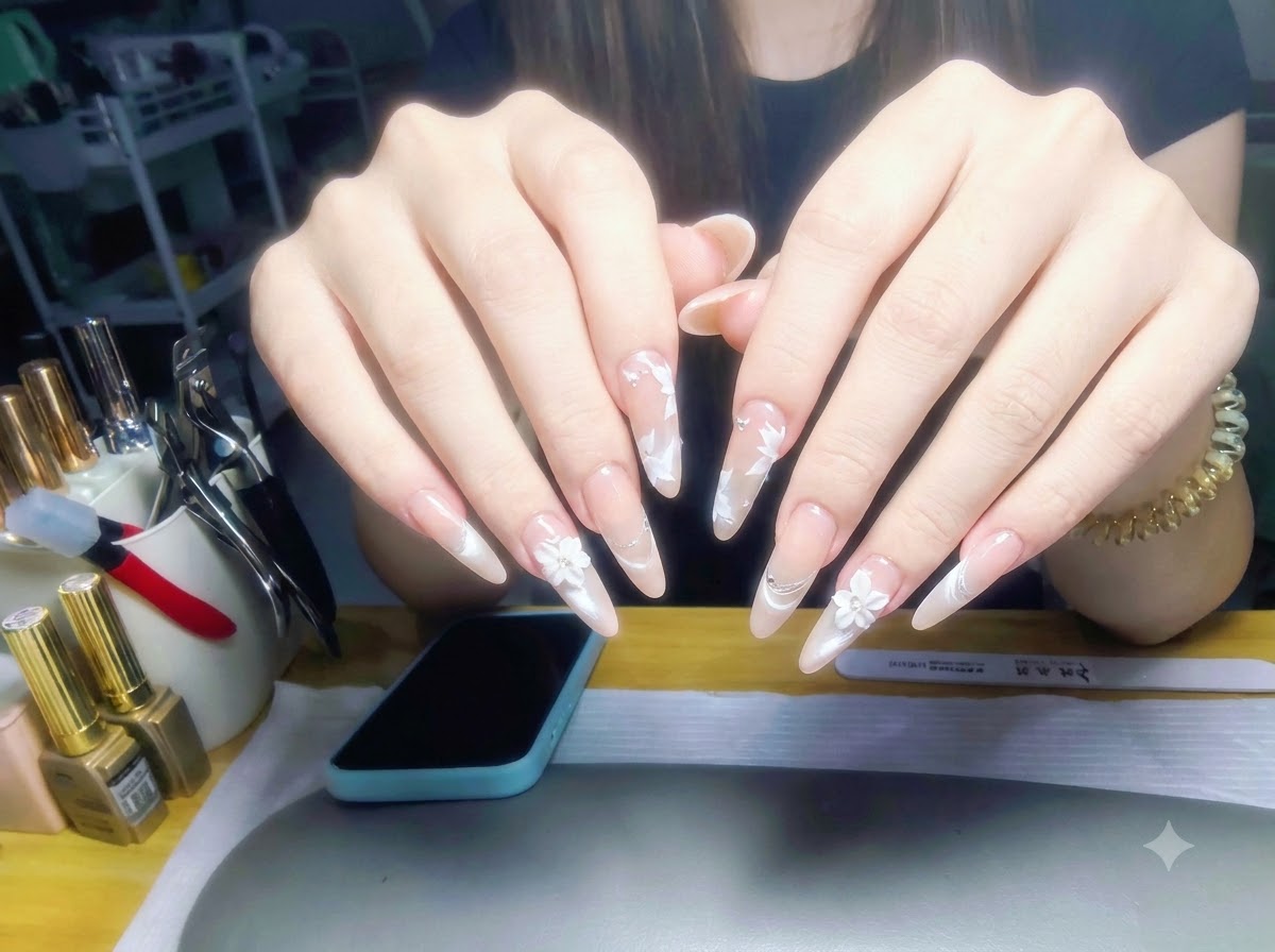 Les deux mains présentées devant un fond de poste de travail, dévoilant une pose complète dongles brillants à dégradé nacré nude et nail art floral blanc.