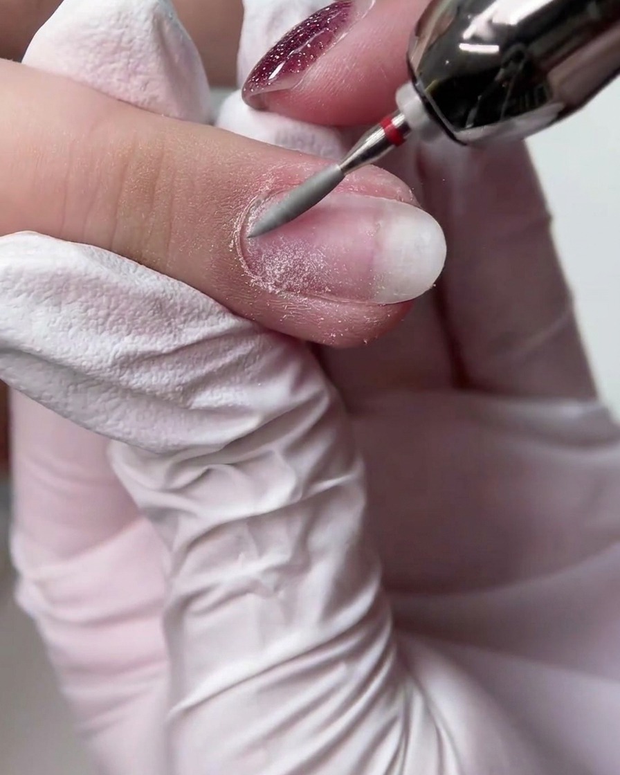 Une main gantée utilisant une ponceuse électrique avec un embout conique long pour lisser la surface dun ongle naturel.