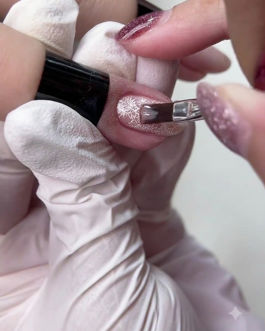 Un petit pinceau à ongles appliquant un vernis gel nude translucide sur un design détaillé dongle en dentelle blanche.