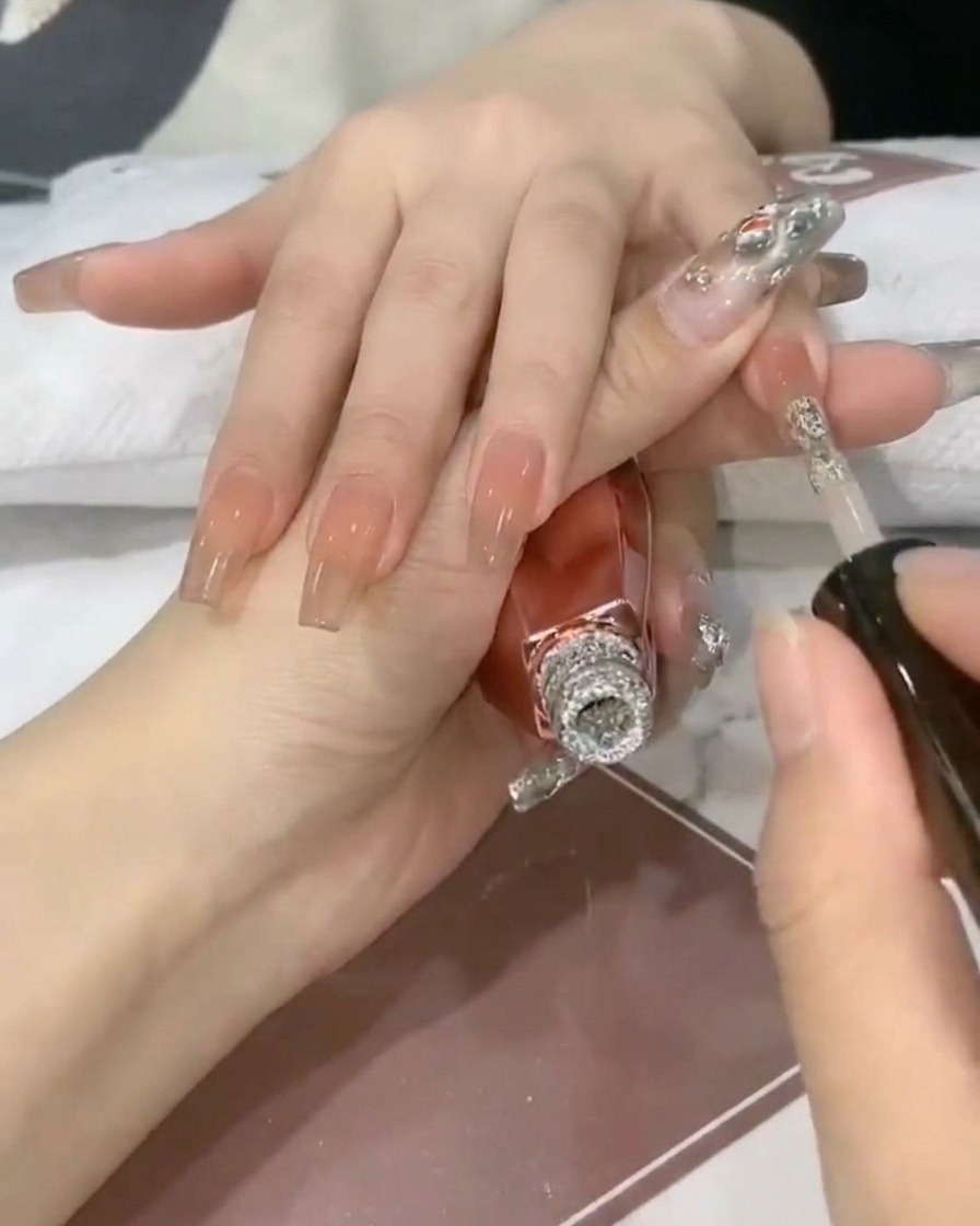 Un pinceau à ongles déposant du gel à paillettes argentées scintillantes sur le bout dun ongle nude translucide.