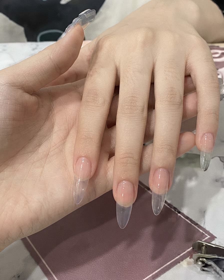 Une main tenant un flacon avec un pinceau appliquant de la colle à ongles transparente sur un ongle préparé.