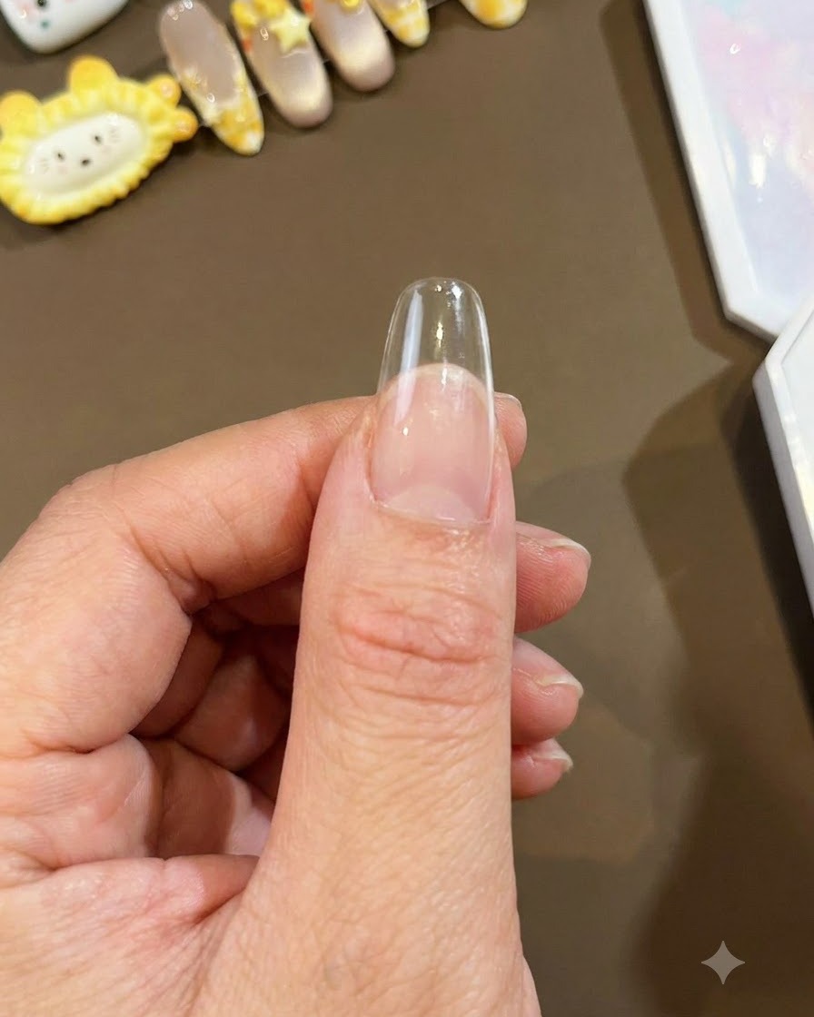 Gros plan dune capsule dongle amande longue et transparente tenue contre un pouce, prête pour lapplication du nail art.