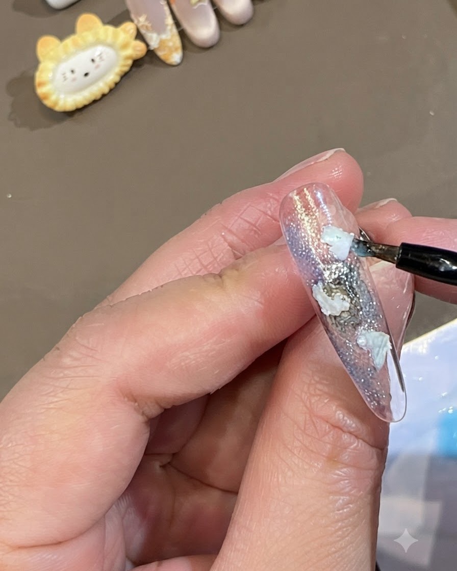 Un pinceau fin déposant des points de gel bleu translucide autour des bords des morceaux de coquillage blancs sur un ongle.