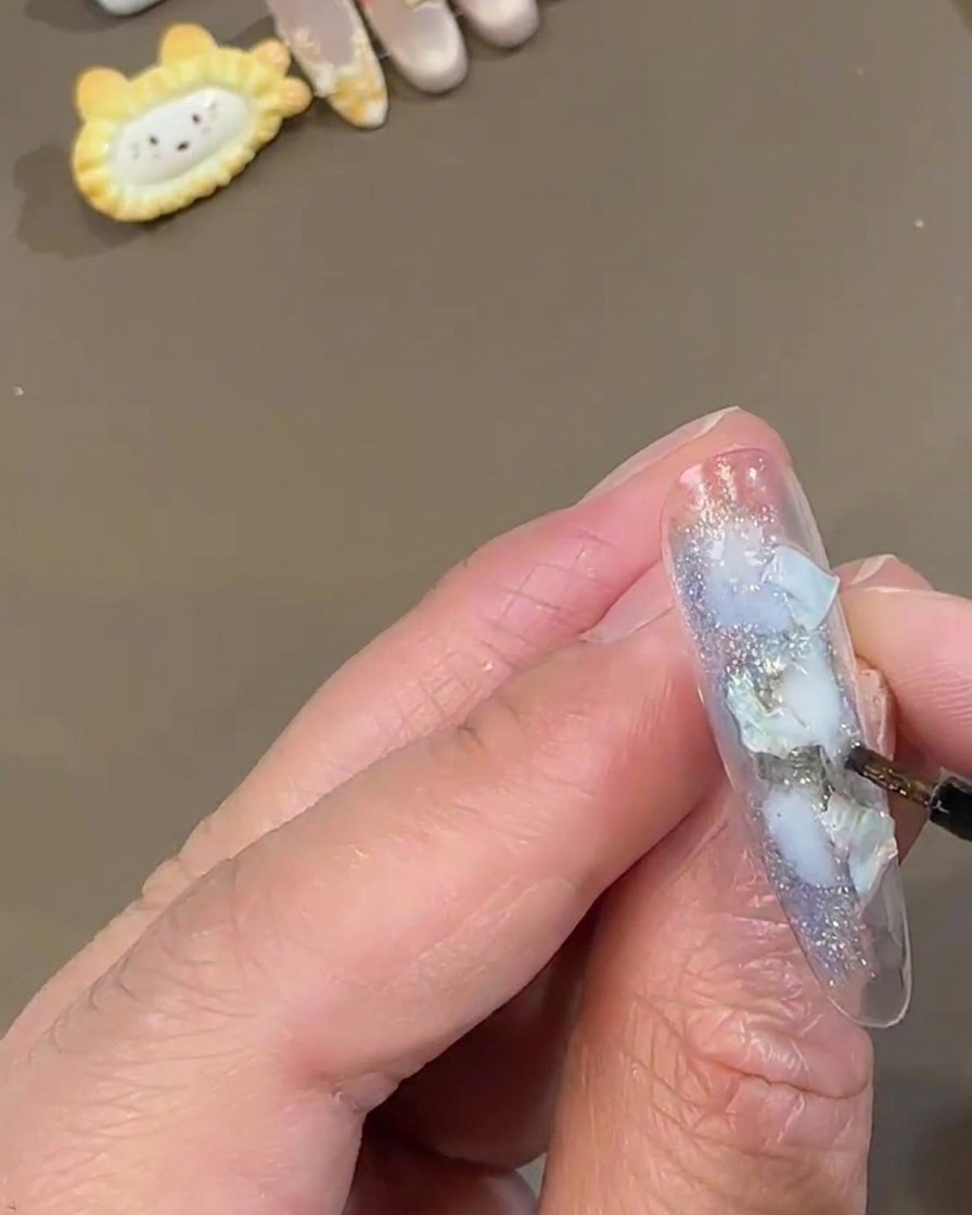 Un pinceau fin pour nail art tamponnant de la poudre de fée blanche scintillante sur un ongle press-on transparent décoré de morceaux de coquillage bleus.