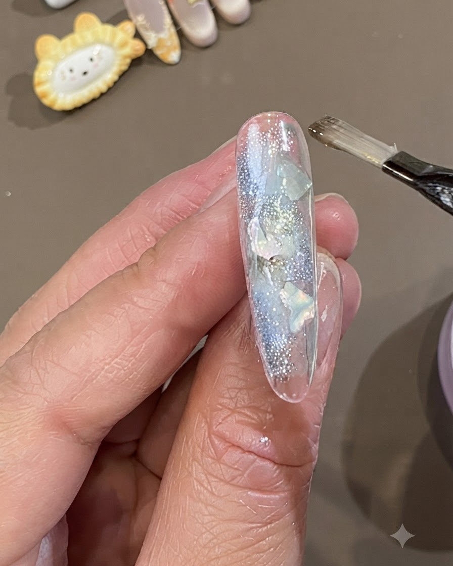 Un pinceau plat à gel appliquant une couche épaisse de gel dencapsulation souple et transparent sur un ongle décoré de morceaux de coquillage et de paillettes.