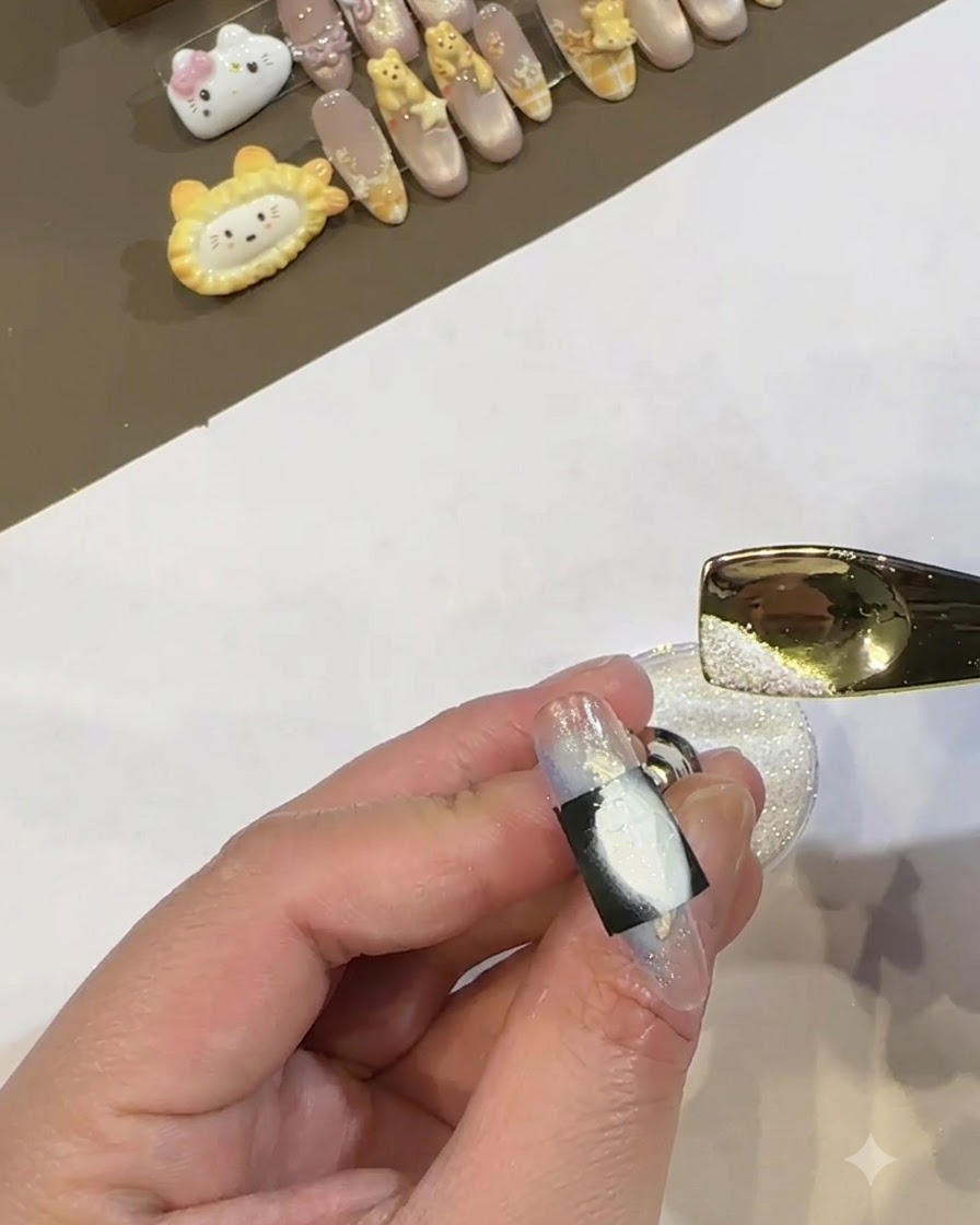 Une cuillère en métal doré versant de la poudre blanche chatoyante sur la zone de gel blanc humide encadrée par un pochoir noir sur un ongle.