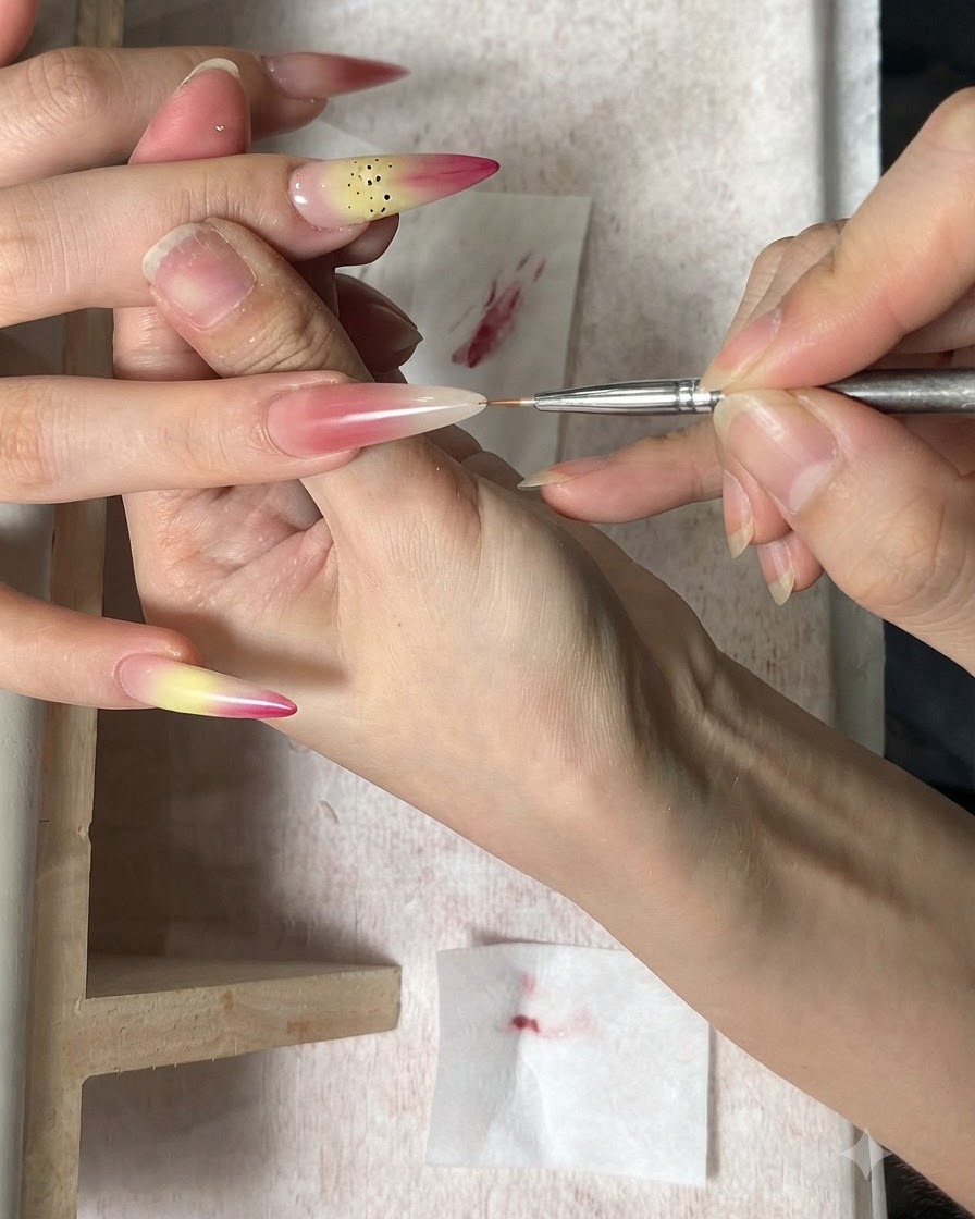 Un pinceau fin pour nail art appliquant des points bruns comme étamines sur un ongle dégradé.
