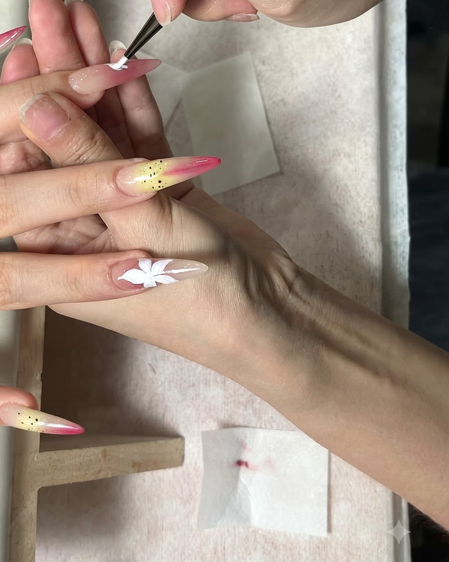 Un pinceau fin peignant une fleur blanche à cinq pétales sur un ongle nude allongé.