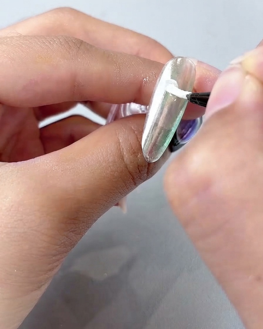 Un pinceau de nail art fin appliquant du gel de peinture blanc en pétales courbés sur une surface dongle chromée argentée.
