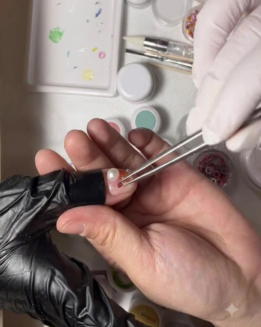 Une main gantée tenant des pinces plaçant une minuscule perle dorée à côté dune perle sur un ongle de pouce décoré.