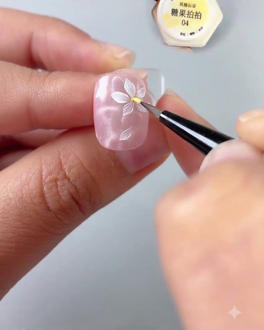 Gros plan dune main tenant un ongle tout en utilisant un pinceau de détail fin pour appliquer une touche de gel jaune au centre dune fleur blanche peinte sur une base dongle rose brillante.