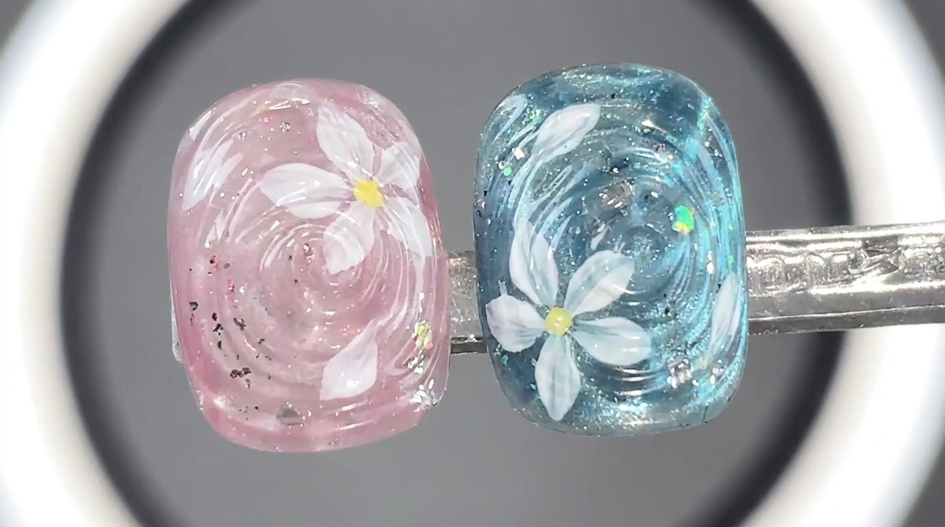 Deux faux-ongles terminés présentant des motifs deau ondulée translucides roses et bleus avec des fleurs blanches et des accents scintillants.