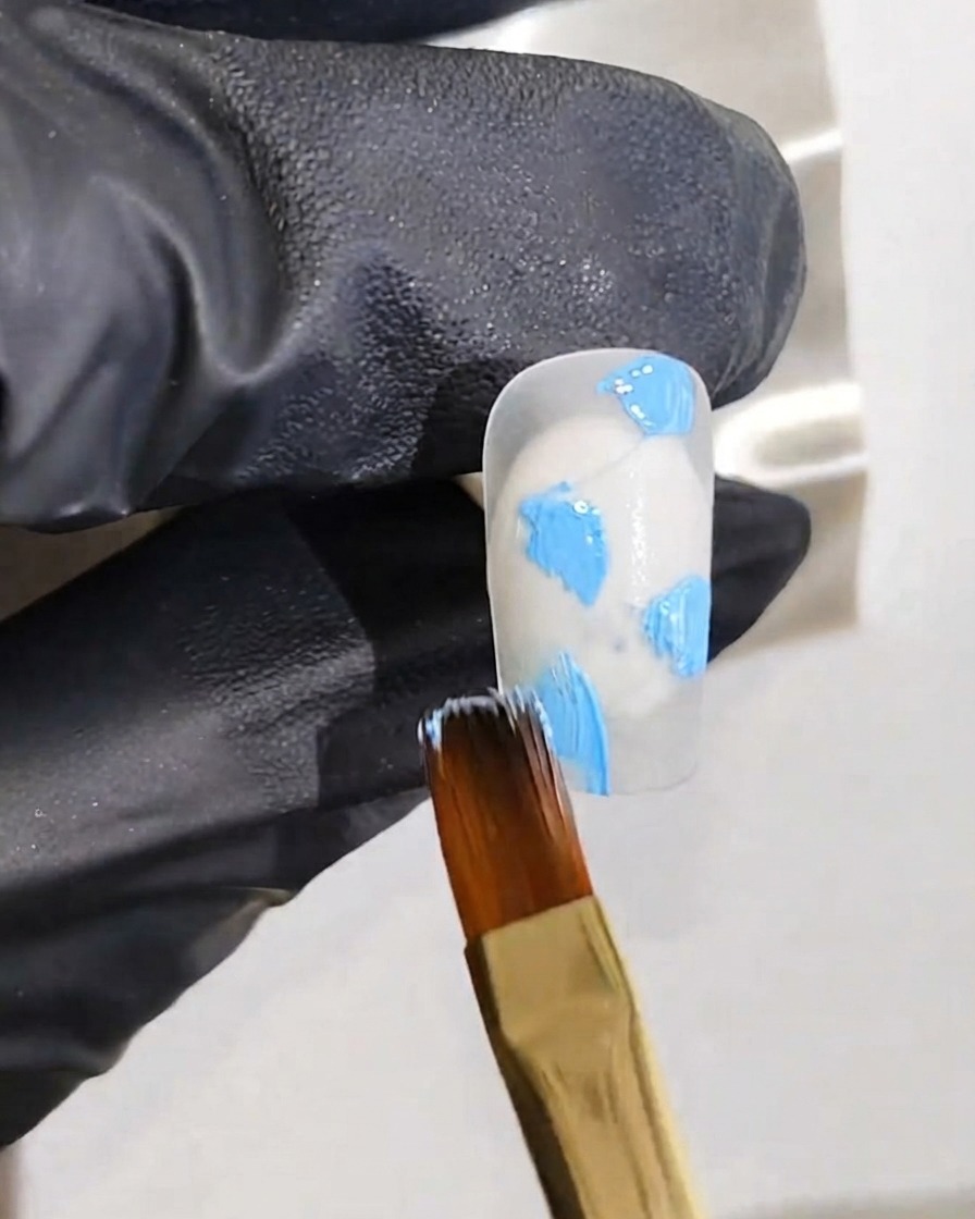 Une main gantée tenant un pinceau fin appliquant des points de gel bleu clair sur une base dongle blanche.