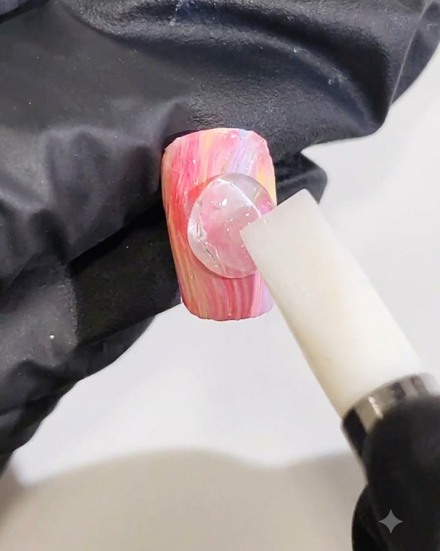 Une main gantée utilisant un outil en silicone blanc pour façonner une goutte de gel 3D en une ligne de vague sur un ongle marbré rose et jaune.