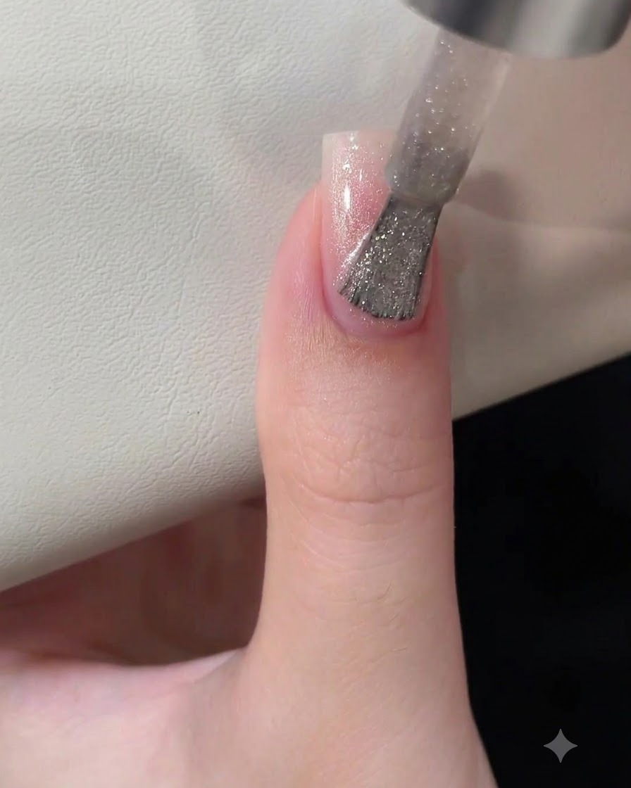 Un pinceau à vernis à ongles appliquant un gel œil-de-chat argenté scintillant sur un ongle en dégradé rose.