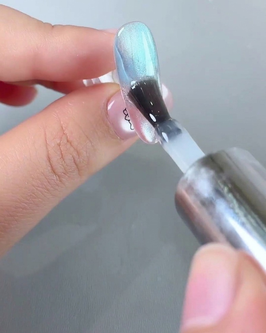 Un pinceau appliquant une couche de gel scellant transparent sur un ongle décoré en ombré rose et bleu.