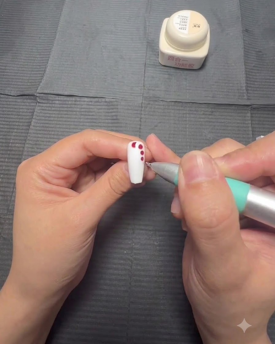 Une main tenant un ongle pendant quun stylet applique des points de gel rouge de plus en plus petits en ligne verticale sur une base blanche.