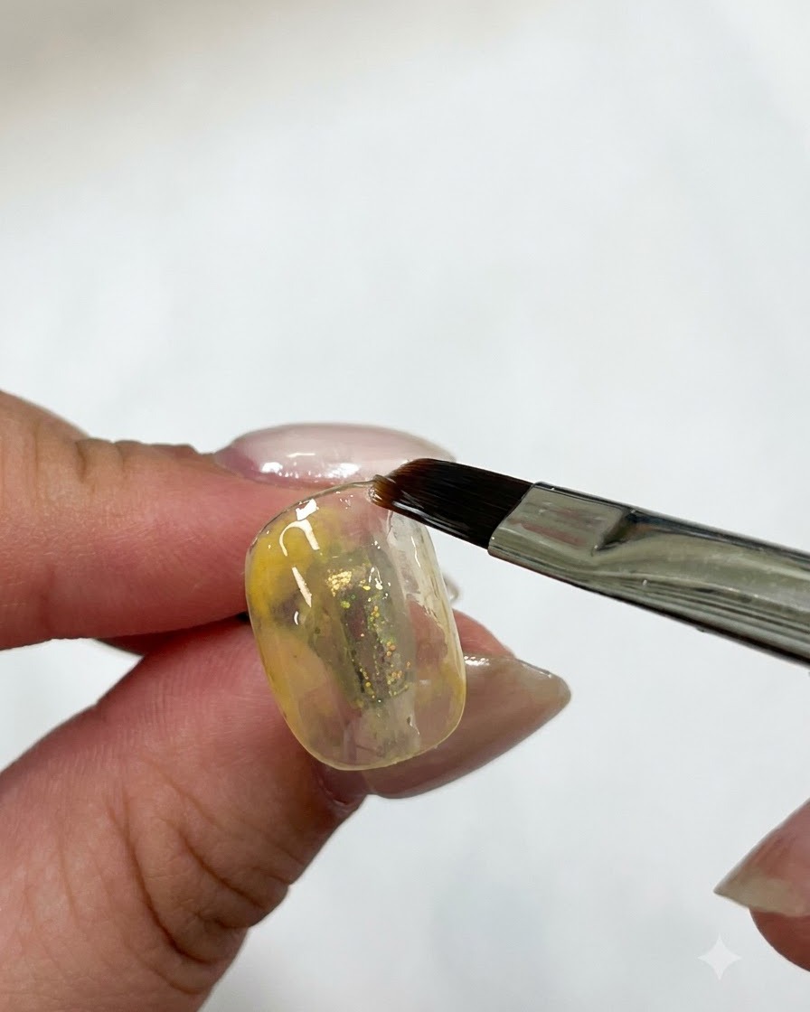 Un pinceau plat à gel lissant la couche de gel jaune sur un ongle sous un éclairage.