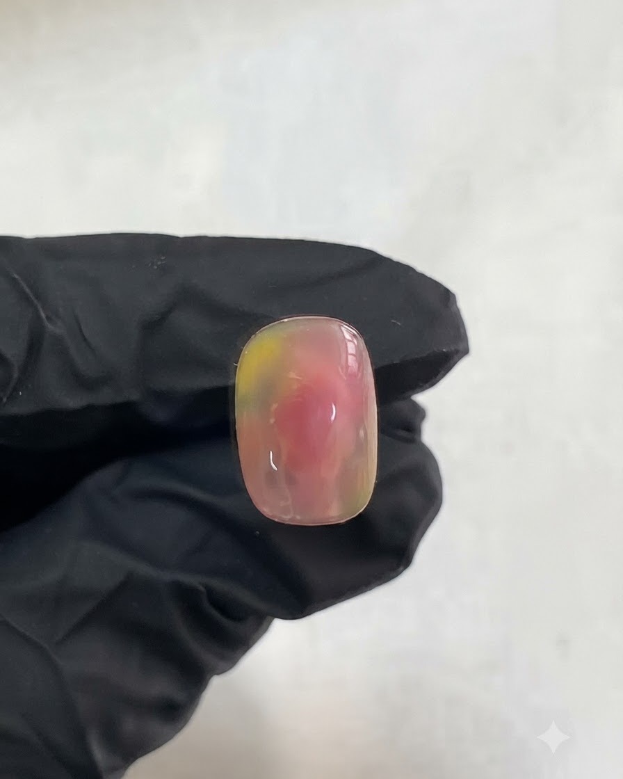 Une main gantée tenant un ongle présentant le design dégradé aquarelle rose et jaune catalysé.