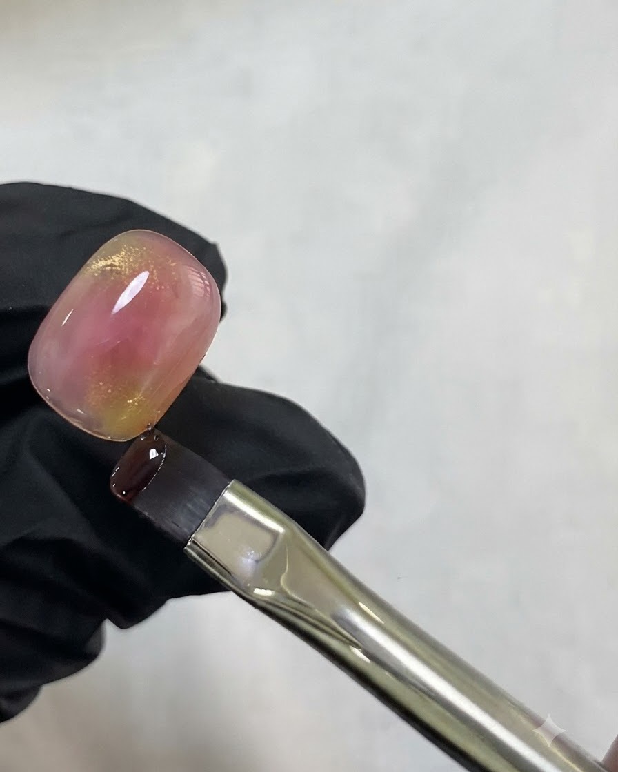 Un pinceau fin retenant une goutte de gel œil de chat doré près du haut dun ongle rose aquarelle.