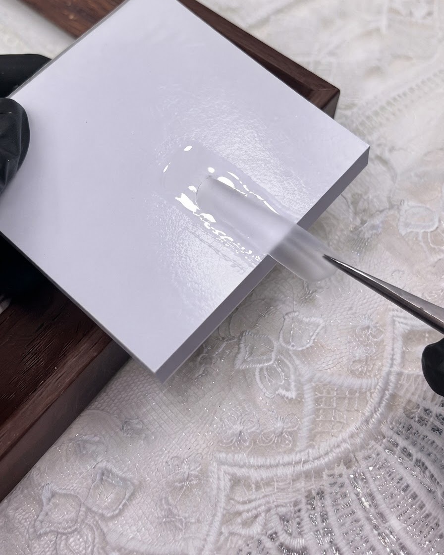Une paire de brucelles plaquant une extension dongle en plastique transparent sur une couche de gel de scellement humide sur une surface blanche.