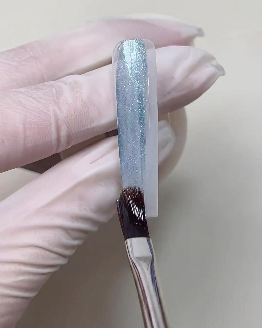 Un pinceau à gel appliquant une ligne épaisse et ondulée de gel cat-eye bleu scintillant au centre dun ongle sur une base blanche.