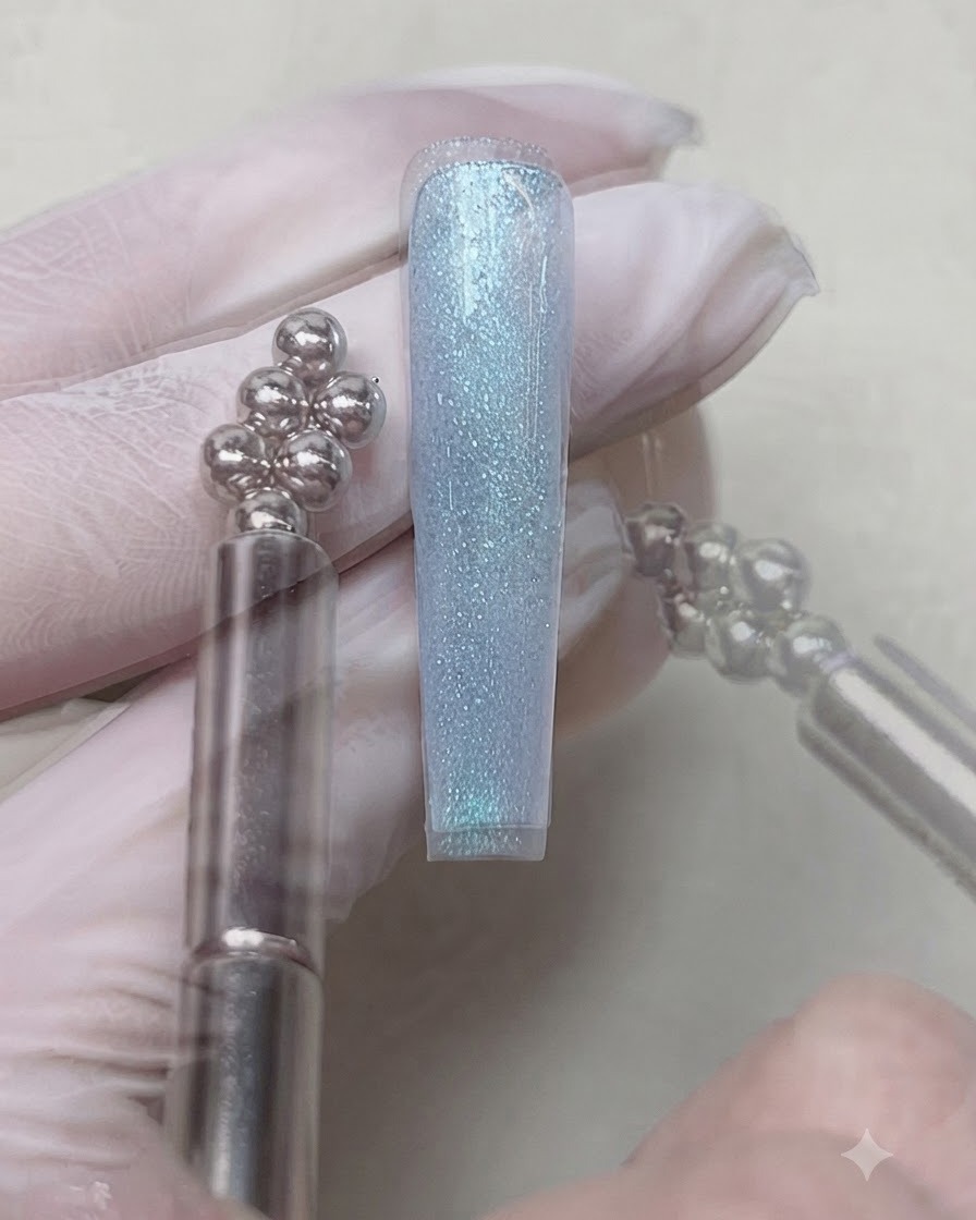 Un outil à aimant sphérique multi-billes tenu près de lextrémité dun ongle pour ajuster le reflet du gel cat-eye bleu.