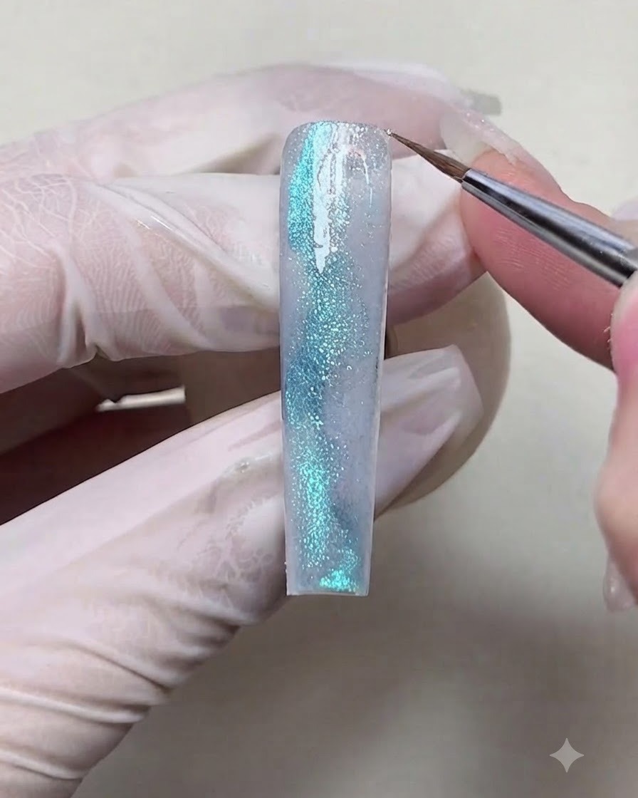 Un pinceau de détail fin appliquant des flocons de paillettes holographiques iridescentes sur certaines zones dun ongle en gel texturé bleu.
