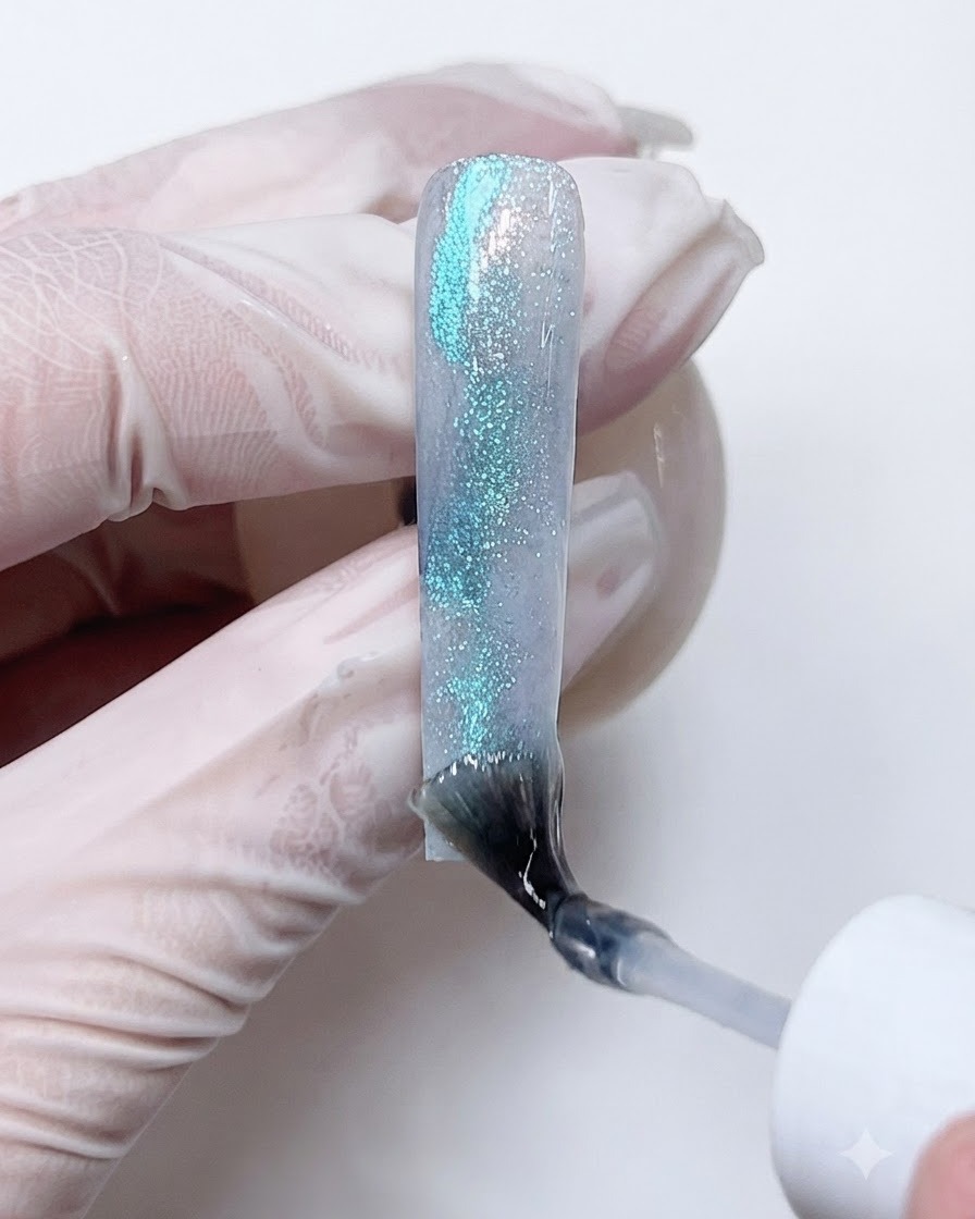 Un pinceau appliquant une couche transparente de gel de construction sur le nail art texturé bleu et pailleté pour encapsuler le design.