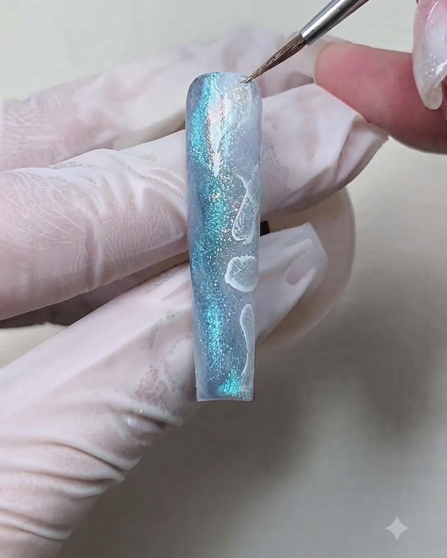 Un pinceau liner fin estompant et plumeant des gouttes dencre blanche pour former une texture décume douce sur un ongle texturé bleu.