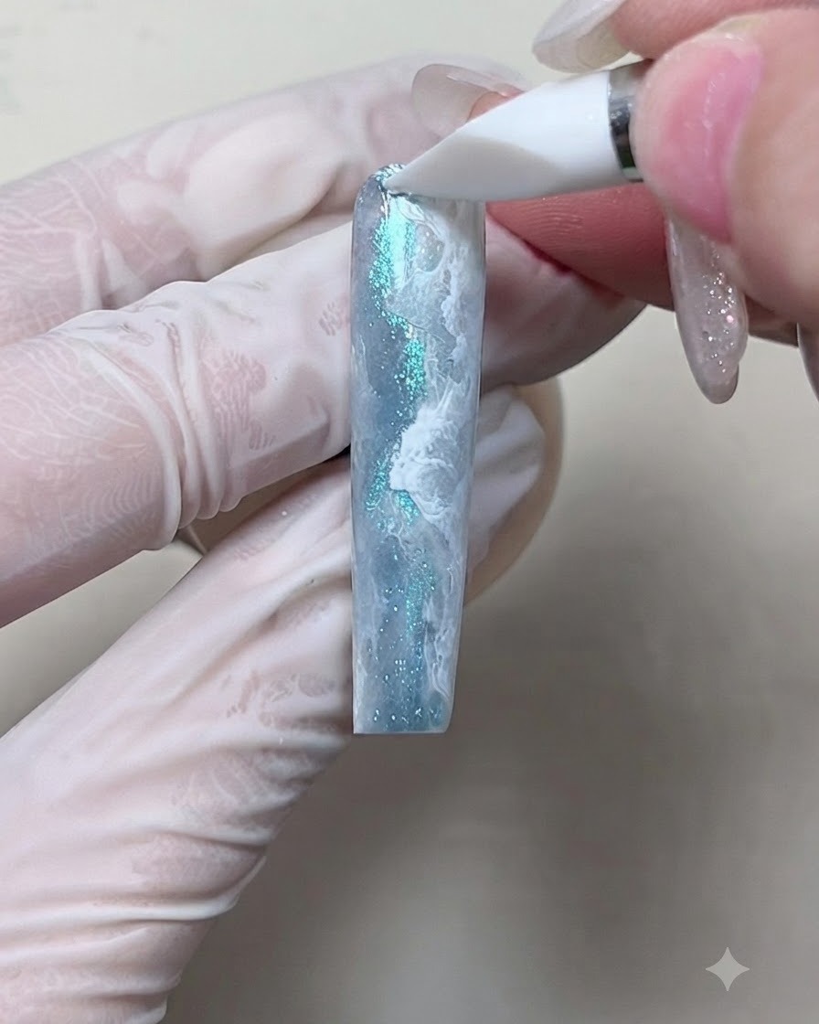 Un outil en silicone appliquant des paillettes bleues scintillantes sur les crêtes blanches dun design de nail art en forme de vague.