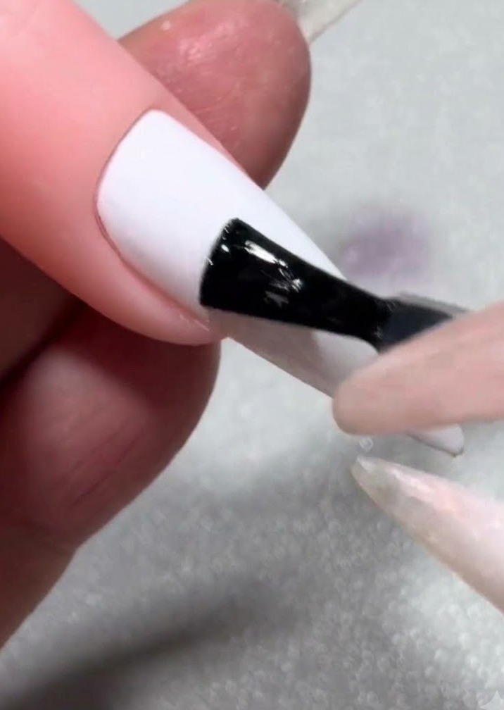 Un pinceau appliquant une couche de blooming gel liquide transparent sur un faux ongle blanc uni.