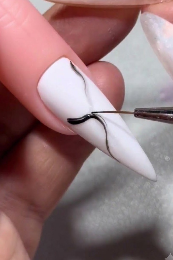 Un pinceau fin ajoutant une touche de couleur foncée concentrée sur une veine de marbre noire en train de sétaler sur un ongle blanc.