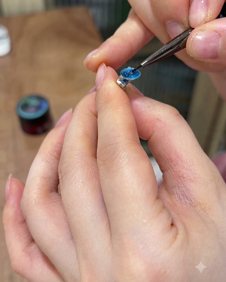 Un pinceau de détail appliquant de la colle à strass solide sur la face arrière plate dune gemme en gel bleu.